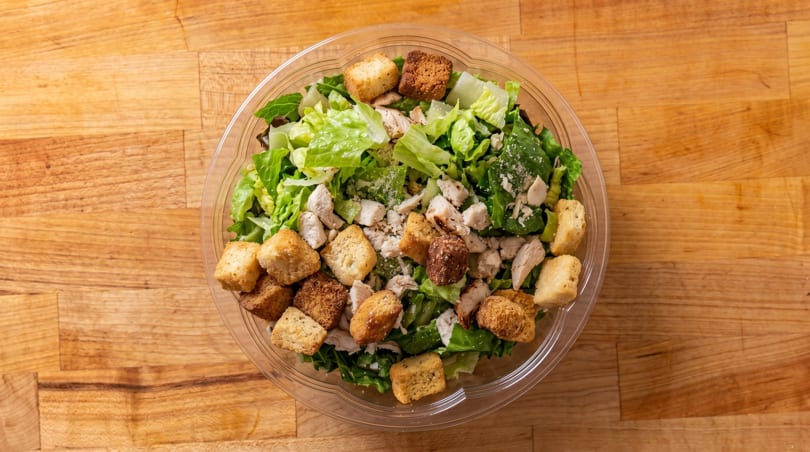 Overhead view of a Caesar salad with chicken, croutons, and parmesan cheese in a plastic bowl.