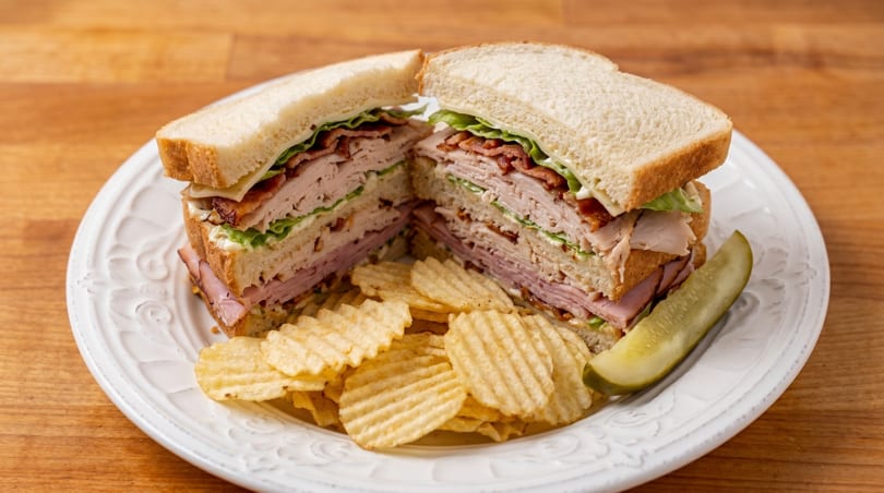 Club sandwich with turkey, ham, bacon, lettuce, and cheese, served with chips and a pickle.