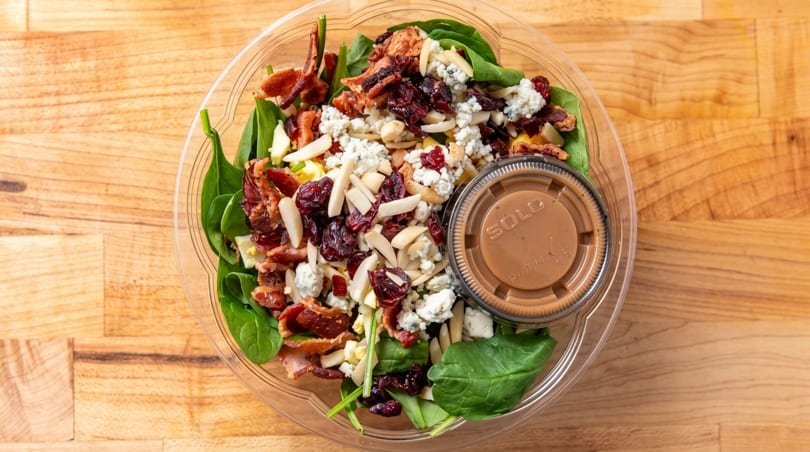 Spinach salad with bacon, blue cheese, cranberries, almonds, and dressing in a clear plastic container.